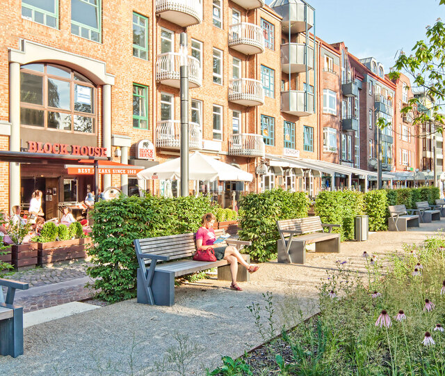 Ahrensburg Blick auf das BLOCK HOUSE Restaurant in Ahrensburg. Im Vordergrund sind ein gepflasterter Weg mit grünen Bäumen und Sitzbänken zu sehen.