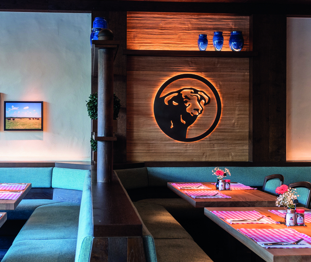 Innenraum des BLOCK HOUSE Restaurants in Köln; mehrere Tischreihen mit Platzsets, Gewürzen und Blumen; das BLOCK HOUSE Bullenlogo wird an einer Holzwand von hinten erleuchtet