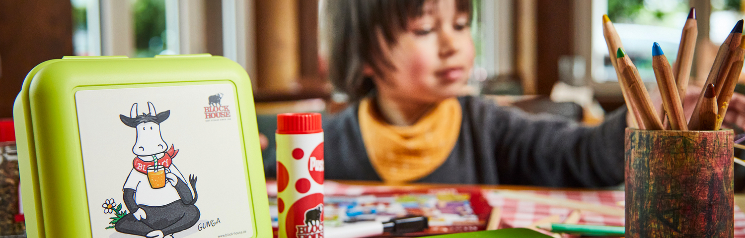 Im Vordergrund sind links eine Blocky Block Brotdose und links ein Becher mit Bunstiften zu sehen. Im Hintergrund malt ein Kind auf einer BLOCK HOUSE Malunterlage