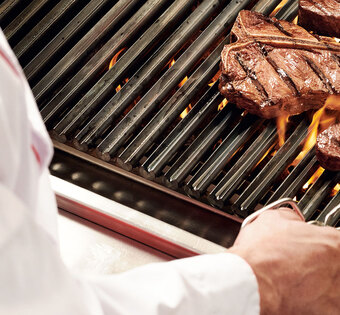 Lübeck Blick über die Schulter eines Grillers, der eine Steakzange in der Hand hält und gerade ein Steak auf einem flammenden Grill wendet.