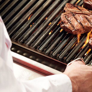 Stuttgart Königstraße Blick über die Schulter eines Grillers, der eine Steakzange in der Hand hält und gerade ein Steak auf einem flammenden Grill wendet.