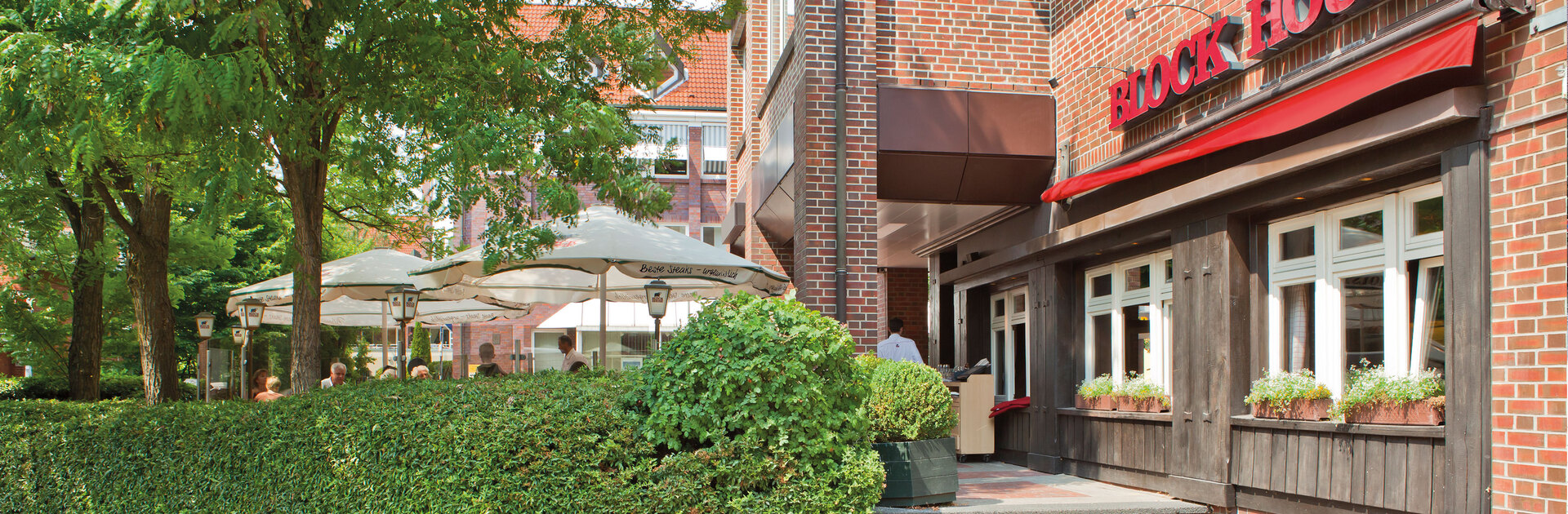Außenfassade des BLOCK HOUSE Restaurants Eidelstedt in Hamburg. Links im Bild ist die Terrasse mit grüner Umrandung und Sonnenschirmen zu sehen. 