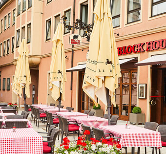 BLOCK HOUSE Restaurant Augsburg Außenansicht des BLOCK HOUSE Restaurants in Augsburg: eine große, hölzerne Fensterfront, davor viele Sitzgelegenheiten auf der Terrasse, die sich direkt auf dem Ulrichsplatz befindet.