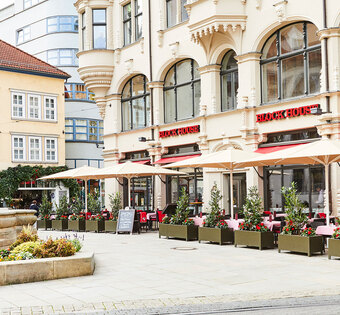 BLOCK HOUSE Restaurant Erfurt Außenansicht des BLOCK HOUSE Restaurants in Erfurt in einem denkmalgeschützten Gebäude . Links ist der alte Angerbrunnen zu sehen. Die Terrasse des Restaurants ist von Pflanzkübeln umsäumt, die Sonnenschirme sind aufgespannt. Das Restaurant hat große Fensterfronten, die den direkten Blick auf den alten Angerbrunnen ermöglichen.