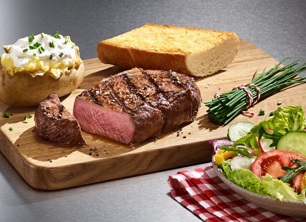 Unsere Steakhouse Klassiker Steak, Baked Potato und BLOCK HOUSE Brot auf einem Holzbrett, serviert mit einem BLOCK HOUSE Salat